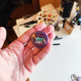 Exhausted Spy illustrated pin button held in hand for size reference.