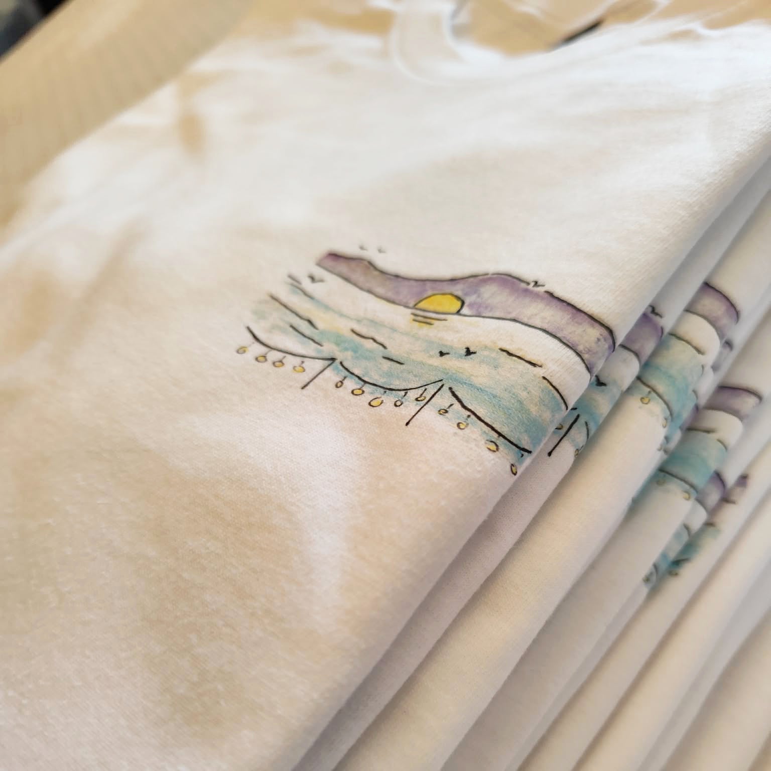 Stack of folded white cotton T-shirts featuring the hand-drawn "Ikarian Dance" illustration.