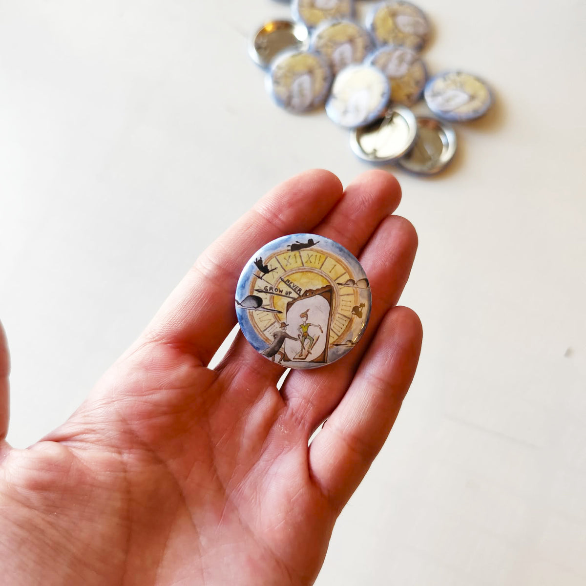 Never Grow Up illustrated pin button held in hand for size reference.