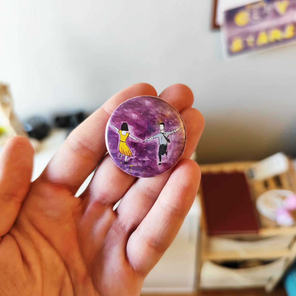 Sunset Dance illustrated pin button held in hand for size reference.